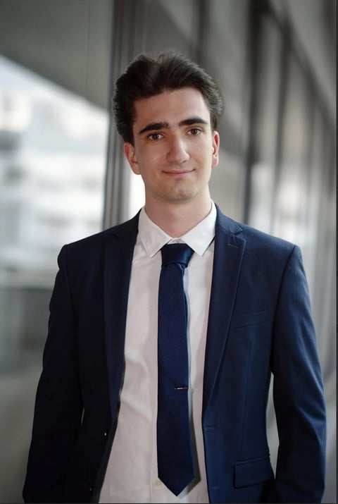 Professional headshot of a man in a dark blue blazer, white shirt, and navy tie against a modern office background